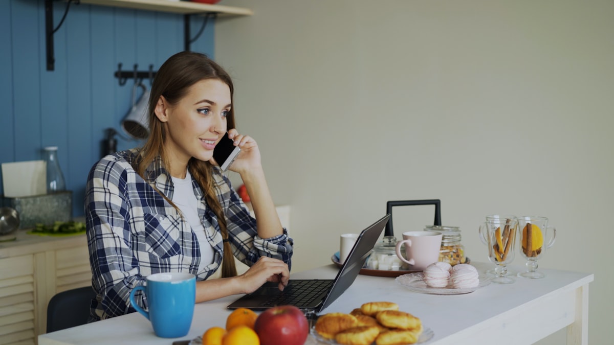 A landlord at her kitchen table, on the phone while checking something on her laptop